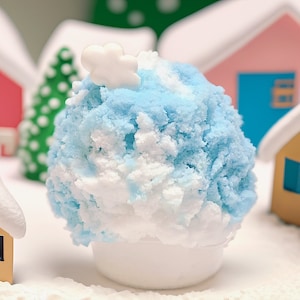 以下が含まれることがあります： 白いボウルに入った、白と青の雪の円錐形。上に白い雲が乗っています。雪の円錐形は、ミニチュアの家とクリスマスツリーに囲まれています。