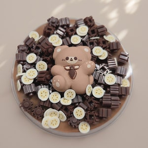 May include: A brown slime base with a light brown teddy bear figure, surrounded by chocolate-colored geometric shapes and banana slices. The slime is in a clear, round container, creating a playful and textured appearance.