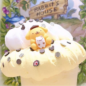 以下が含まれることがあります： ホイップクリームとチョコレートスプリンクルで飾られた黄色と白のデザート。デザートの上に、茶色の帽子をかぶった小さな黄色い犬の置物が置かれています。デザートの後ろの看板には「Rabbit's House」と書かれています。