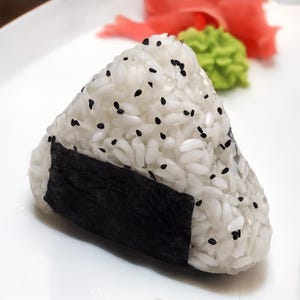 May include: A triangular onigiri rice ball with black sesame seeds, wrapped partially in a sheet of nori seaweed. The onigiri sits on a white plate, with pickled ginger and wasabi in the background. A Japanese cuisine staple.
