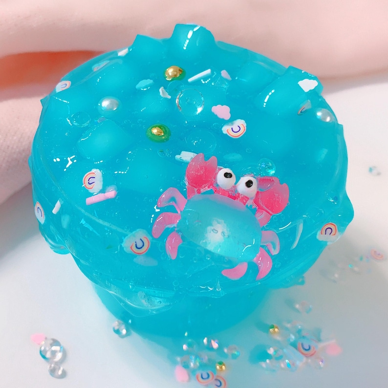 May include: Blue slime with a pink and white crab, white and gold sprinkles, and iridescent glitter.