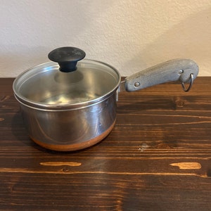 May include: A silver saucepan with a copper bottom and a glass lid. The saucepan has a black knob on the lid and a gray handle with a metal ring.
