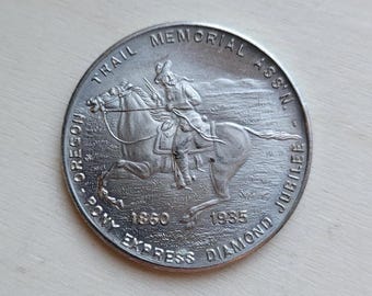 Op de afbeelding: Een zilveren munt met een ruiter te paard, met de tekst "OREGON PONY EXPRESS DIAMOND JUBILEE 1860-1935 TRAIL MEMORIAL ASSN." De munt heeft een gedetailleerd reliëfontwerp en een cirkelvorm.