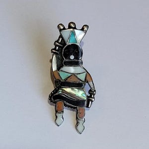 May include: A silver-toned pin featuring a stylized Native American figure. The figure has a black head, a white and turquoise headdress, and is adorned with abalone shell and other colorful inlays. The figure is holding a staff.