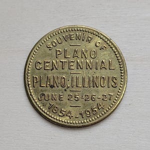 May include: A round, brass-colored souvenir coin. The coin reads "SOUVENIR OF PLANO CENTENNIAL PLANO, ILLINOIS JUNE 25-26-27 1854-1954" in a circular pattern around the edge. The coin has a beaded border.