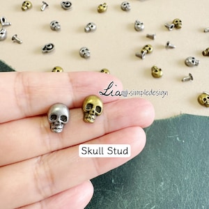 Peut inclure: Gros plan sur des clous en forme de crâne argentés et dorés. Les clous sont petits, avec des détails de crâne. L'image montre également plusieurs autres clous en forme de crâne éparpillés. Le texte sur l'image indique "Skull Stud".