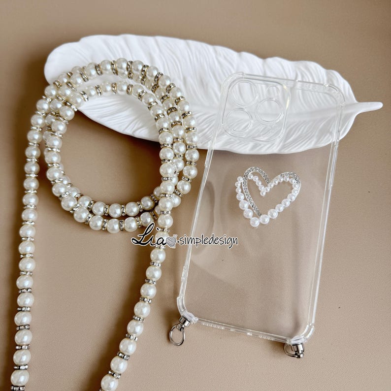 May include: Clear phone case with a pearl and rhinestone heart design. A pearl and rhinestone phone strap is draped next to the case. The text "Lia simpledesign" is visible on the bottom left of the image. The background is a neutral beige color.