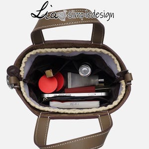 May include: A brown leather tote bag with a beige lining. The bag is open and the inside is filled with various items, including a red lipstick, a small bottle of hand sanitiser, and a white phone.