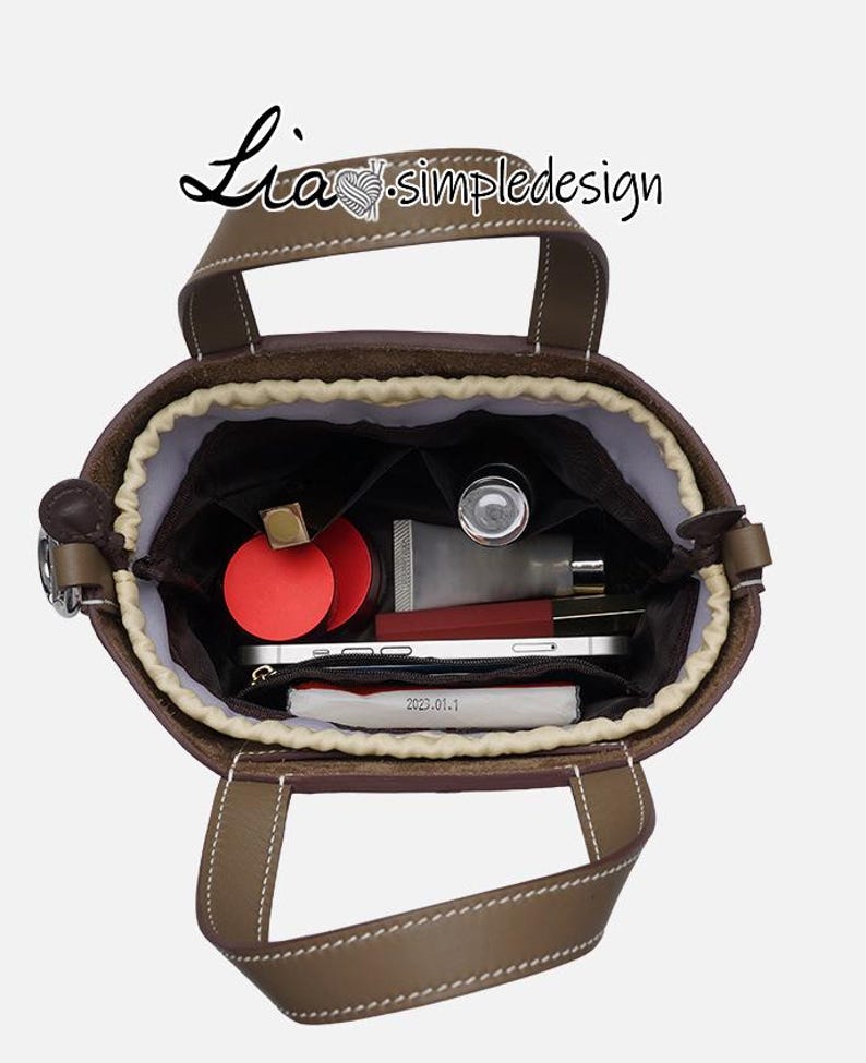 May include: A brown leather tote bag with a beige lining. The bag is open and the inside is filled with various items, including a red lipstick, a small bottle of hand sanitiser, and a white phone.