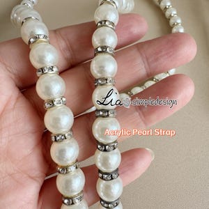 May include: Close-up of an acrylic pearl strap with large, round, white pearls. The pearls are interspersed with silver-colored rhinestone spacers. The text "Lia simpledesign" and "Acrylic Pearl Strap" are visible.