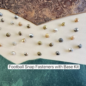Puede incluir: Una colección de broches a presión con forma de balón de fútbol en varios acabados metálicos, incluyendo plata, oro y metal de cañón. Los broches están sobre una superficie de cuero. El texto dice "Football Snap Fasteners with Base Kit".