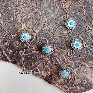 May include: Six silver metal conchos with turquoise centres are displayed on a brown leather surface. The leather has a floral embossed pattern. Each concho features a starburst design around the turquoise centre. The leather has a distressed appearance.