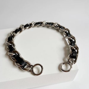 Peut inclure: Un bracelet en cha&icirc;ne m&eacute;tallique argent&eacute;e avec un cordon en cuir ou similicuir noir entrelac&eacute; dans les maillons. Le bracelet est dot&eacute; d'un fermoir &agrave; ressort circulaire &agrave; chaque extr&eacute;mit&eacute;, permettant de relier les deux c&ocirc;t&eacute;s.