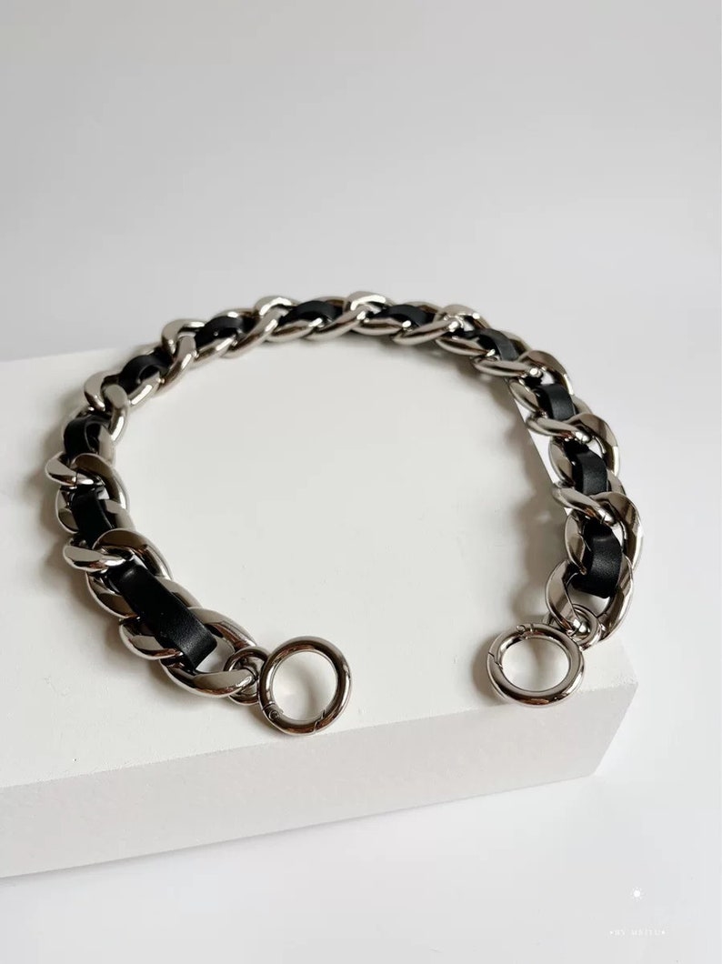 Peut inclure: Un bracelet en cha&icirc;ne m&eacute;tallique argent&eacute;e avec un cordon en cuir ou similicuir noir entrelac&eacute; dans les maillons. Le bracelet est dot&eacute; d'un fermoir &agrave; ressort circulaire &agrave; chaque extr&eacute;mit&eacute;, permettant de relier les deux c&ocirc;t&eacute;s.