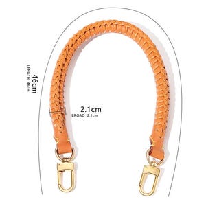 May include: Tan leather handbag handle with a braided design and gold-coloured clasps. The handle is 46 cm long and 2.1 cm wide. The handle is arched. The clasps are a shiny gold colour.