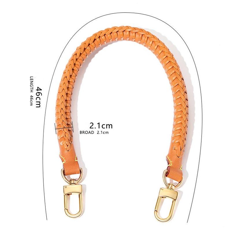 May include: Tan leather handbag handle with a braided design and gold-coloured clasps. The handle is 46 cm long and 2.1 cm wide. The handle is arched. The clasps are a shiny gold colour.