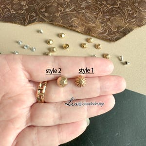 Puede incluir: Dos estilos de tachuelas de metal dorado, una con un diseño de estrella y la otra con un diseño facetado. El texto "style 1" y "style 2" está escrito debajo de las tachuelas.