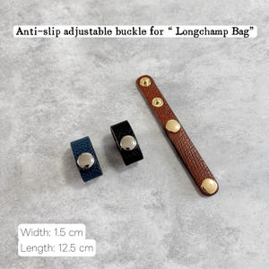 May include: An anti-slip adjustable buckle for a Longchamp bag. The image shows three buckles: one brown with gold studs, one black, and one navy blue. The width is 1.5 cm and the length is 12.5 cm.