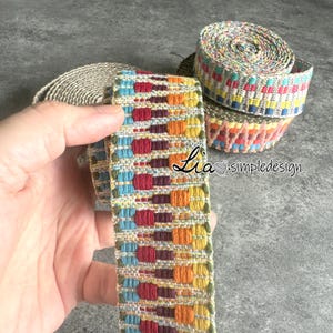 May include: A close-up of colorful woven trim with a repeating pattern of red, orange, yellow, blue, and green. The trim is rolled up and held in a hand.