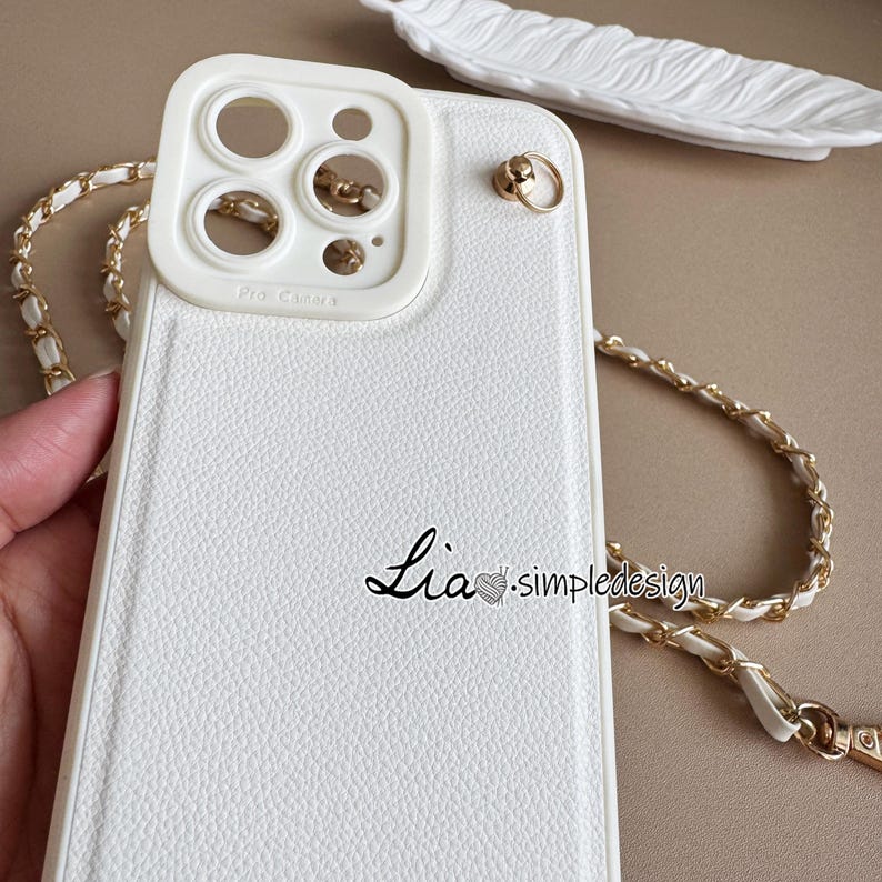 May include: White phone case with a textured finish, featuring a gold chain strap. The case has a camera cutout and the text "Lia. simpledesign" in black script. A decorative feather-shaped dish is in the background.