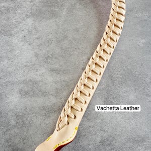 May include: A close-up of a cream-coloured leather strap with a braided design. The strap is labelled "Vachetta Leather" and features yellow and red stitching at the end. The strap is on a grey background.