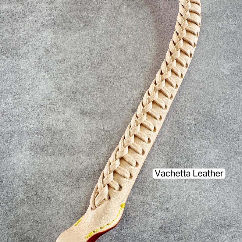 May include: A close-up of a cream-coloured leather strap with a braided design. The strap is labelled "Vachetta Leather" and features yellow and red stitching at the end. The strap is on a grey background.