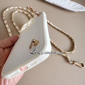 May include: White phone case with a gold chain strap. The case has a gold ring holder and the text "Lia.simpledesign". The chain is white with gold accents. The phone case is made of a smooth material.