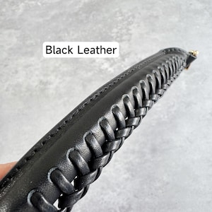 May include: Close-up of a black leather strap with a braided design. The strap has a stitched edge and a gold-coloured metal clasp. The text "Black Leather" is displayed in a white rectangle.
