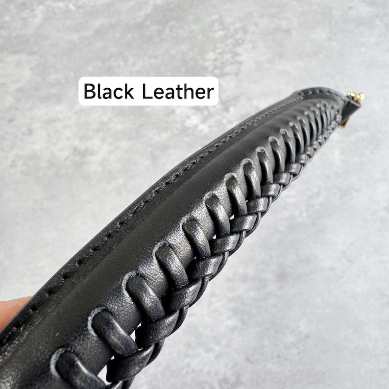 May include: Close-up of a black leather strap with a braided design. The strap has a stitched edge and a gold-coloured metal clasp. The text "Black Leather" is displayed in a white rectangle.