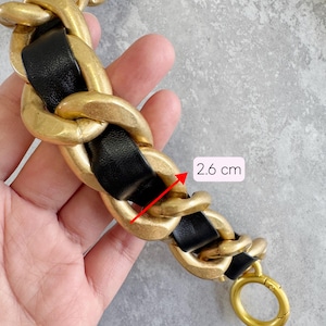 Peut inclure: Une bandouli&egrave;re d&eacute;corative compos&eacute;e de grands maillons en m&eacute;tal dor&eacute; mat entrelac&eacute;s d'une bande de cuir noir lisse. Une fl&egrave;che rouge indique une largeur de maillon de 2,6 cm. La cha&icirc;ne se termine par un fermoir annulaire en m&eacute;tal dor&eacute;.