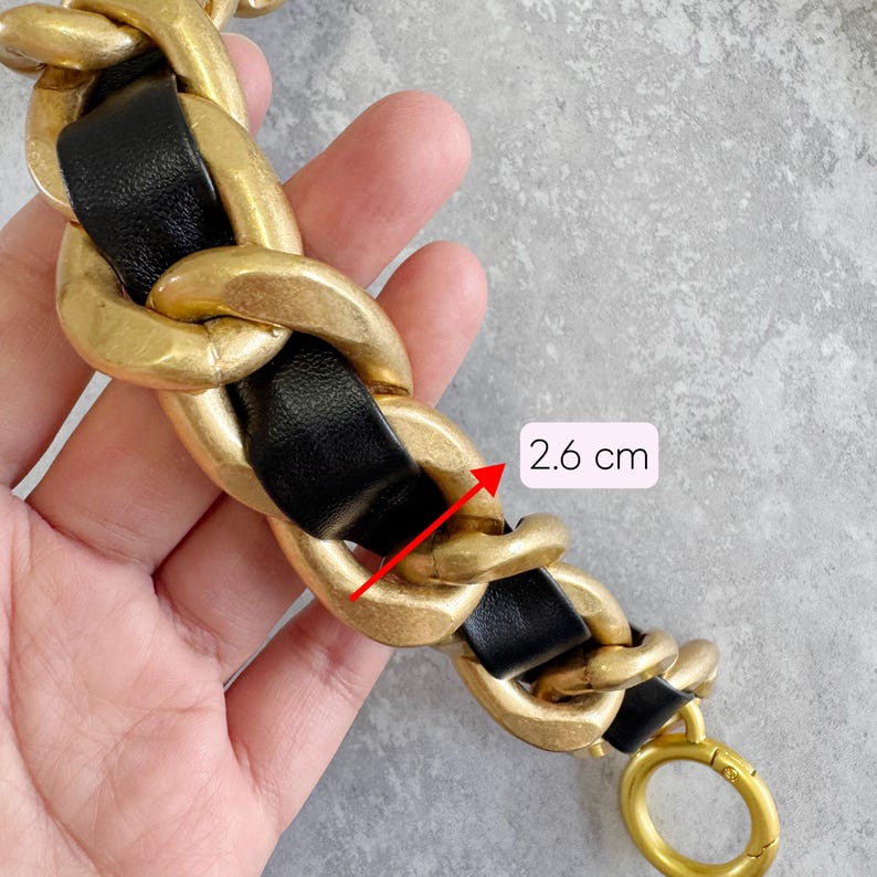 Peut inclure: Une bandouli&egrave;re d&eacute;corative compos&eacute;e de grands maillons en m&eacute;tal dor&eacute; mat entrelac&eacute;s d'une bande de cuir noir lisse. Une fl&egrave;che rouge indique une largeur de maillon de 2,6 cm. La cha&icirc;ne se termine par un fermoir annulaire en m&eacute;tal dor&eacute;.