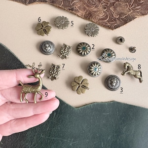 May include: Assortment of metal charms in various shapes and designs, including a deer, owl, clover, lion, and flowers. The charms are in shades of gold and green.
