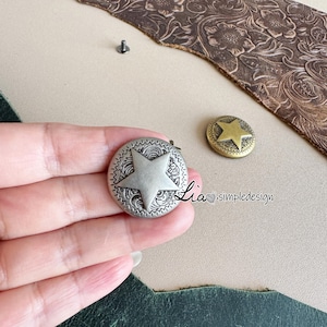 May include: Two metal star-shaped buttons are shown. One is silver with a star in the center and a decorative border. The other is gold-coloured with a star design. These buttons are likely for clothing or accessories.