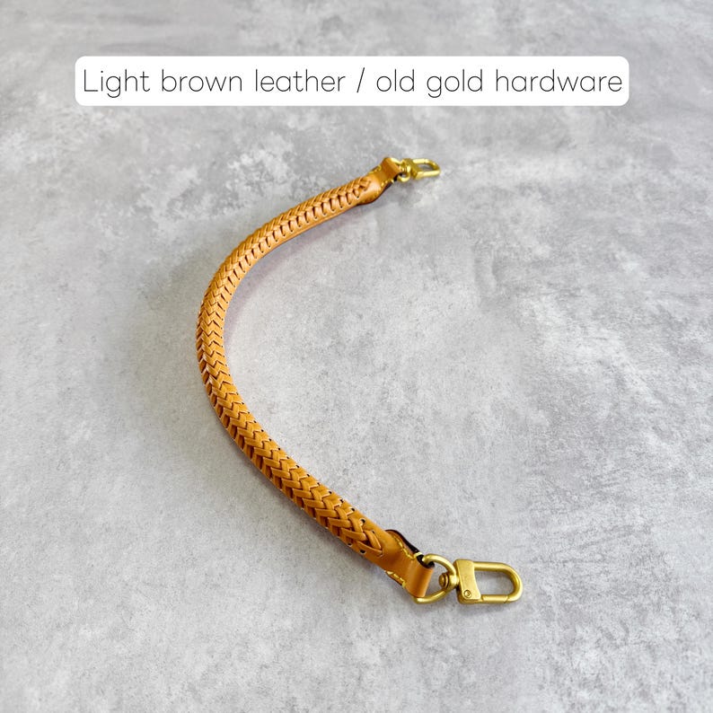 May include: A light brown leather handbag strap with a braided design. The strap has old gold hardware, including a clasp at each end. The text "Light brown leather / old gold hardware" is visible in the image.