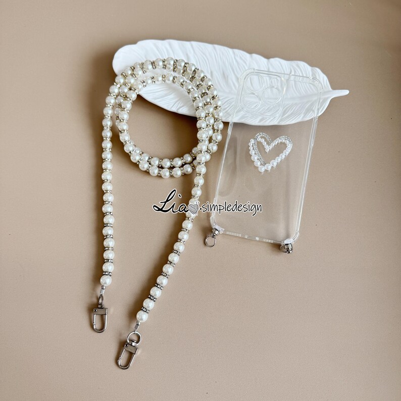May include: A clear phone case with a pearl heart design and a pearl strap. The strap has silver-colored clasps. The text "Lia simpledesign" is printed on the case.
