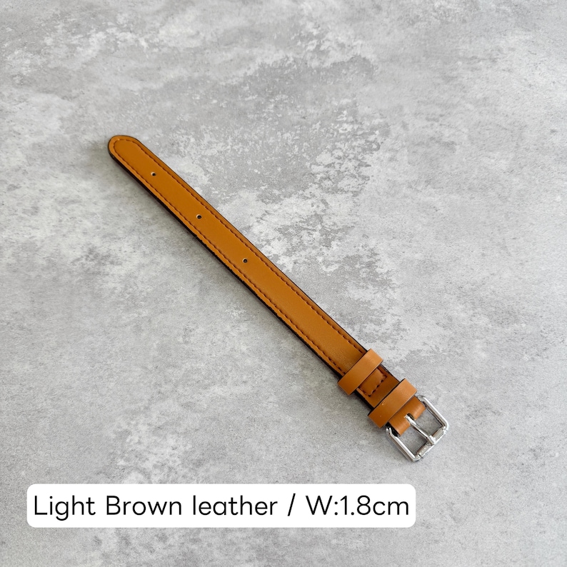 May include: A light brown leather watch strap featuring a silver-toned metal buckle and two matching leather keepers. The strap has a smooth finish with visible stitching along the edges and includes several adjustment holes. The width is 1.8 cm.