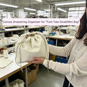 Könnte beinhalten: Ein cremefarbener Canvas-Kordelzug-Organizer für eine kleine oder Mini-Tragetasche. Er hat einen Kordelzugverschluss mit braunen Lederakzenten und eine Vordertasche. Der Text "Canvas Drawstring Organizer for Font Tote Small/Mini Bag" ist sichtbar.