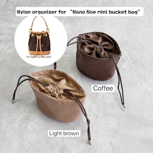 Peut inclure: Deux organisateurs en nylon pour mini sac seau Nano Noe, l'un marron café et l'autre marron clair. Chaque organisateur dispose d'une fermeture à cordon avec des liens marron foncé et des embouts métalliques. Le texte indique Nylon organizer for Nano Noe mini bucket bag.