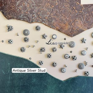 Puede incluir: Una variedad de tachuelas de plata antigua en diversas formas, incluyendo estrellas, cruces y diseños circulares. La imagen incluye el texto "Antique Silver Stud". Las tachuelas están sobre una superficie de cuero texturizado.
