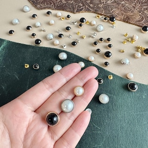 May include: A selection of decorative buttons in various sizes, featuring pearl, black, and gold details. The buttons are arranged on a green and tan surface. Some buttons are held in a hand, displaying the different styles.