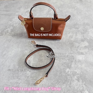 May include: A brown handbag with a matching flap and handle, with a gold-tone charm. A separate black and brown strap with gold-tone clasps is also shown. The text "THE BAG IS NOT INCLUDED" is visible. The strap length is 65 cm.