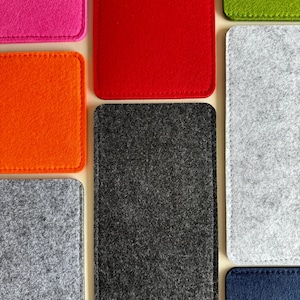May include: An assortment of colorful felt sleeves in various shades, including pink, red, orange, green, gray, black, and blue. Each sleeve is rectangular with rounded corners and stitched edges, showcasing a minimalist design.