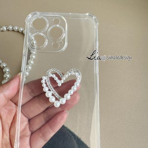 May include: Clear phone case with a heart-shaped embellishment. The heart is outlined with small, clear crystals and surrounded by white pearls. A pearl strap is visible. The text "Lia simpledesign" is printed on the case.