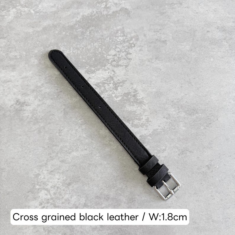 May include: A cross-grained black leather watch strap featuring a silver-toned metal buckle and two matching leather keepers. The strap has a width of 1.8 cm as indicated by the text. The surface of the leather displays a fine, textured pattern.
