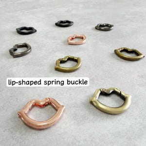Puede incluir: Varias hebillas de resorte con forma de labio en bronce, oro rosa y negro. Las hebillas están dispuestas sobre una superficie gris. El texto "lip-shaped spring buckle" se muestra en un rectángulo blanco.