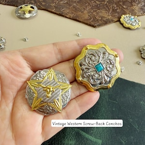 Pode incluir: Dois conchos de metal redondos com fechos de parafuso. Um apresenta uma estrela dourada com uma cabeça de boi no centro sobre uma base prateada gravada. O outro é um design floral prateado com uma borda festonada dourada e uma pedra turquesa quadrada.