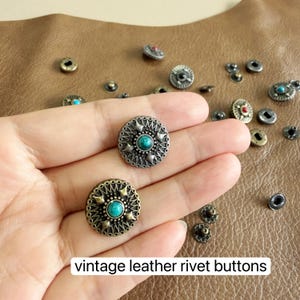 May include: A collection of round metal rivet buttons featuring intricate embossed patterns and central turquoise-coloured stones. The buttons have a metallic finish and are scattered alongside two buttons held in the palm of a hand.