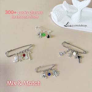 May include: Silver safety pins with various charms, including a dolphin, a guitar, a cross, and a snowflake. The text "300+ Lucky charms to choose from" and "Mix & Match" are visible.