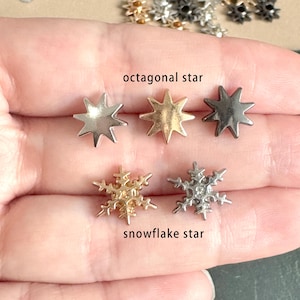 Puede incluir: Primer plano de una mano sosteniendo seis adornos de metal en forma de estrella. Dos estrellas octogonales doradas, dos estrellas octogonales plateadas y dos estrellas de copo de nieve plateadas.