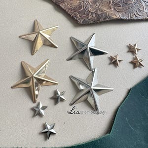 May include: Assortment of gold and silver star-shaped embellishments. The stars are various sizes and are arranged on a white surface. The text "Lia - simpledesign" is visible in the bottom right corner of the image.